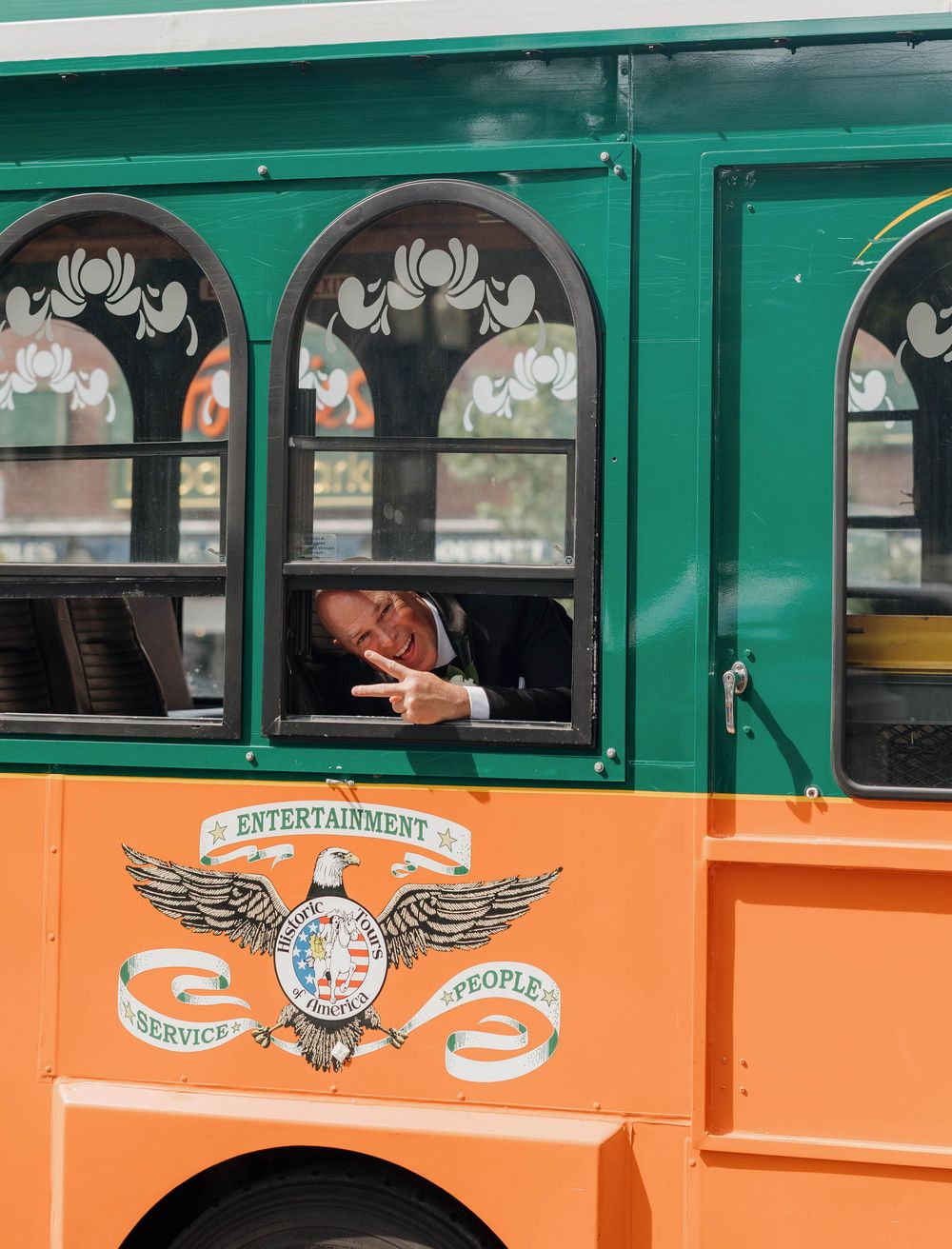 A vintage-style orange and green trolley tour bus with decorative arched windows and an entertainment logo on the side.