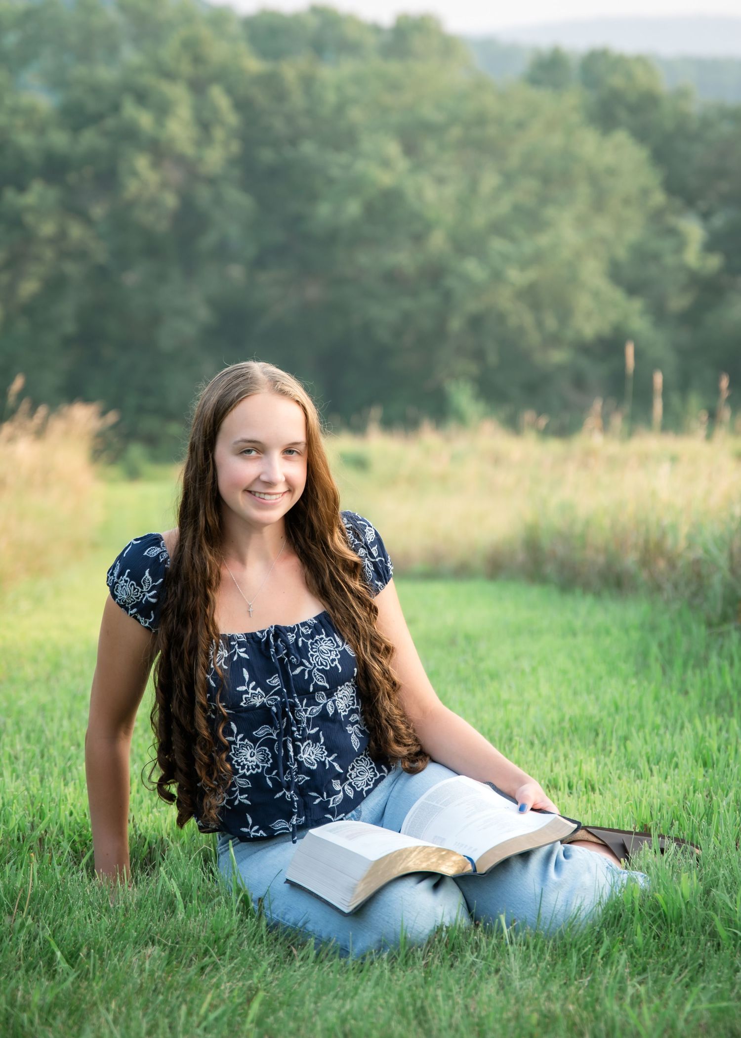 A person in a navy floral top sits reading a book in a sunlit grassy meadow on a summer afternoon.