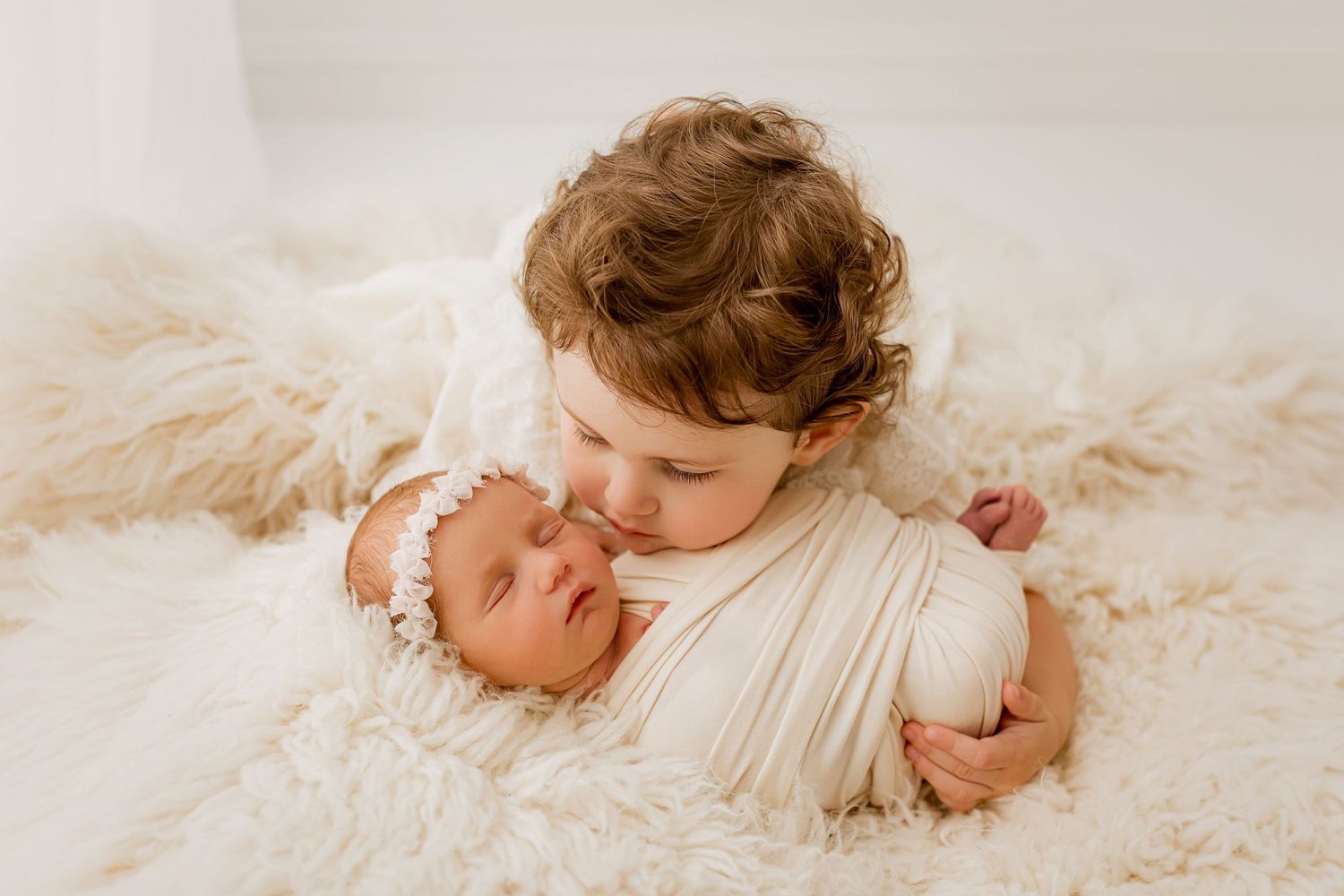 Toddler sister kissing sleeping newborn baby in cream wrap and white headband on fur blanket during sibling portrait session