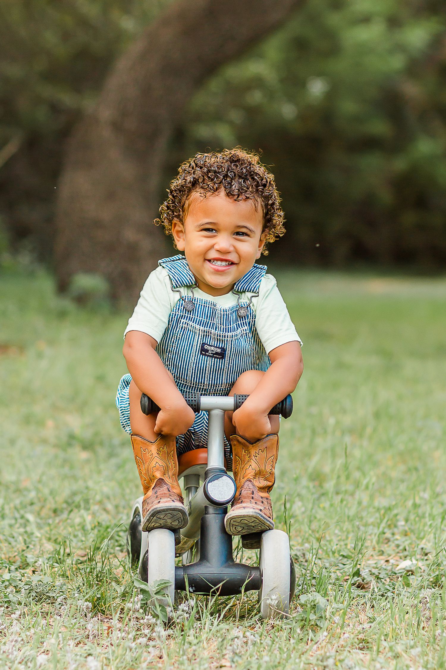 A toddler in denim overalls sits on a white tricycle in a grassy outdoor setting.