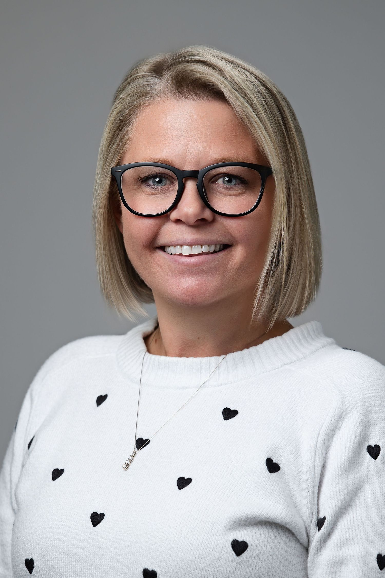 A person wearing glasses and a white sweater with small black heart patterns smiles at the camera in a studio setting.