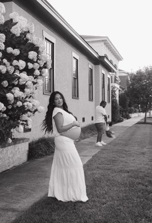 A black and white photograph shows someone in a flowing white dress posing outdoors next to a house with climbing flowers.