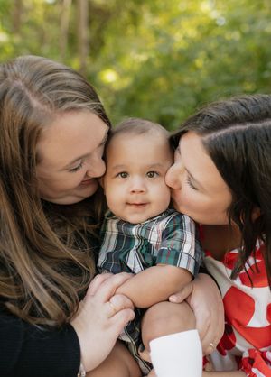 A baby in a plaid shirt smiles while being kissed on both cheeks in an outdoor setting with soft bokeh lighting.