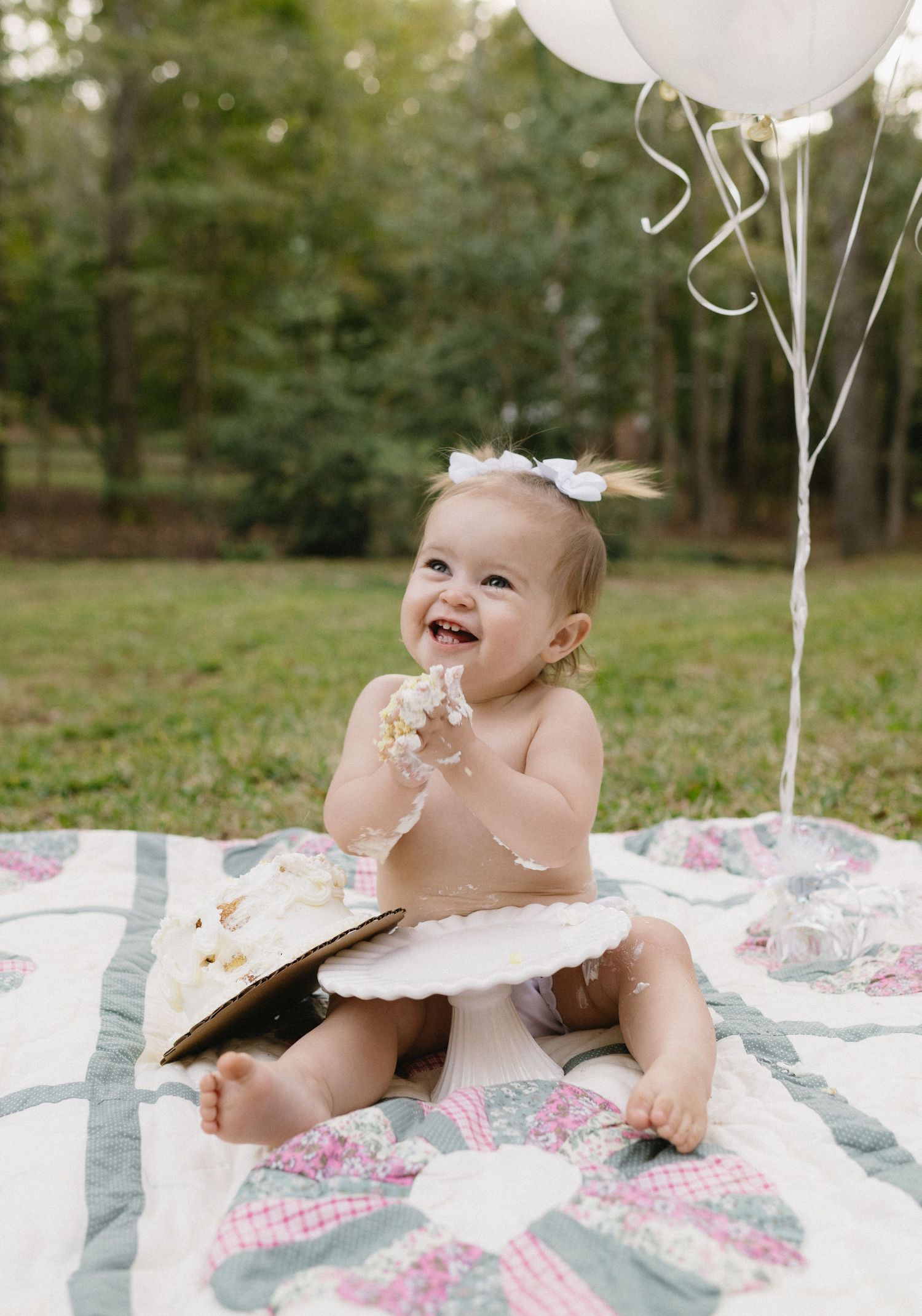 Baby wearing white bow sits on floral blanket with white balloons during cake smash photo session.