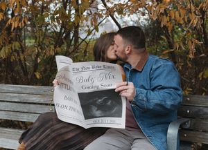 A couple sits together on a wooden bench in autumn reading a newspaper in a park setting.