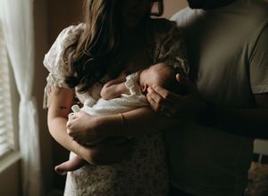 A newborn baby swaddled in white fabric is held lovingly during an intimate indoor photo session.