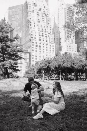 A family relaxes on the grass in Central Park with towering Manhattan apartment buildings visible in the background.