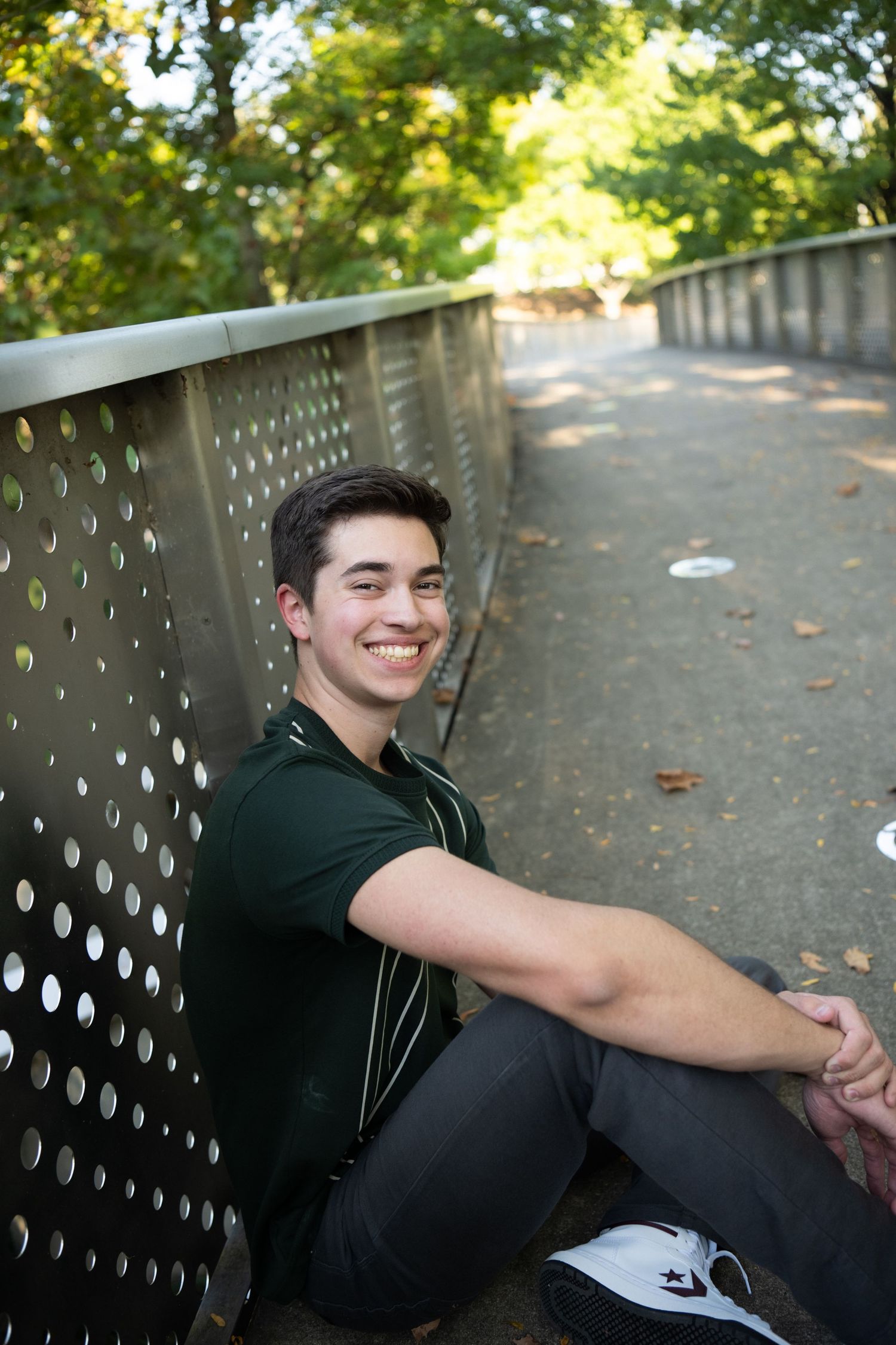 Person in dark clothing sits on wooden bench in sunlit park with perforated metal backrest.