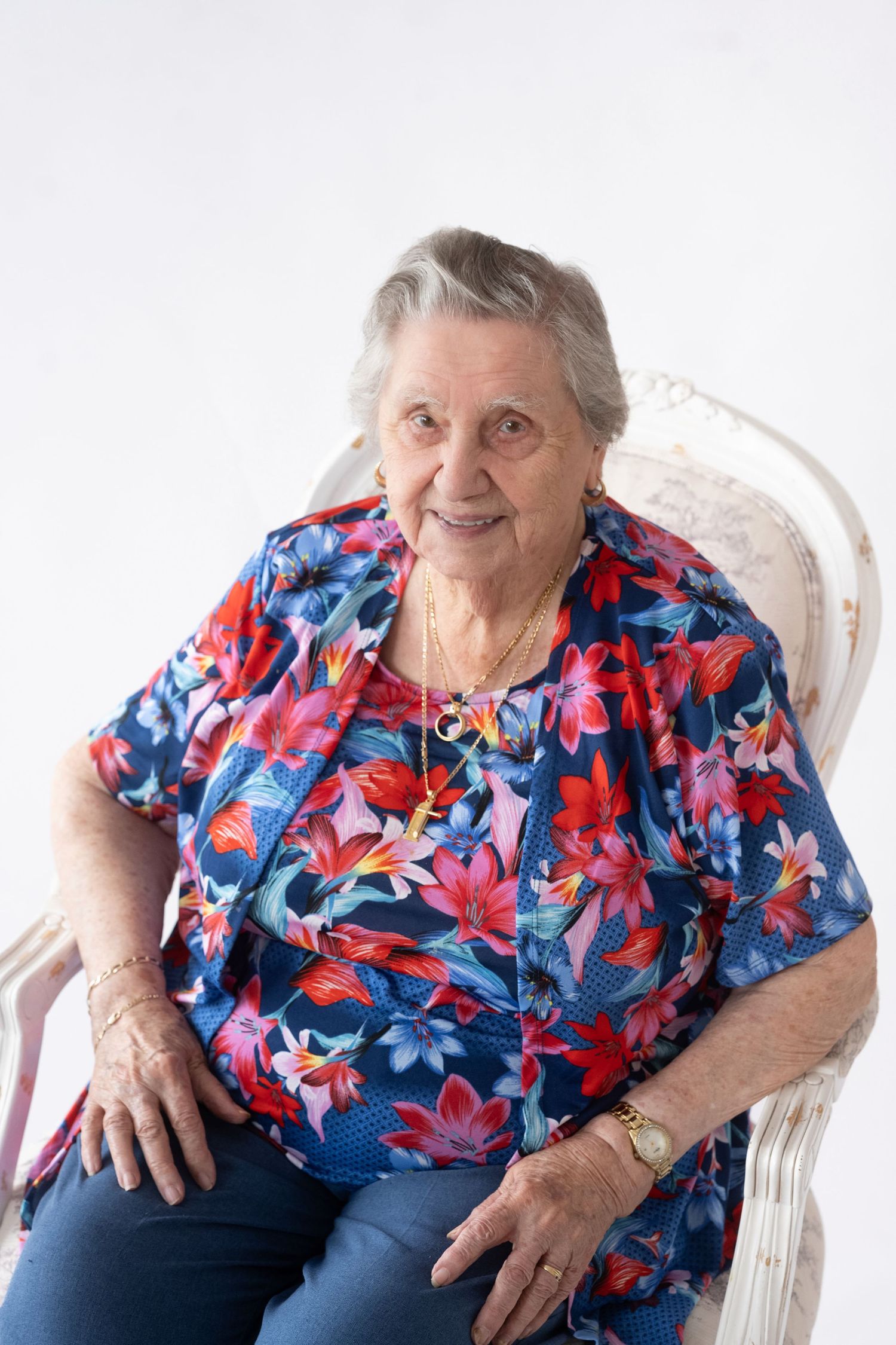 Elderly woman in colorful floral blouse smiles while sitting in white chair, shown from three different angles.