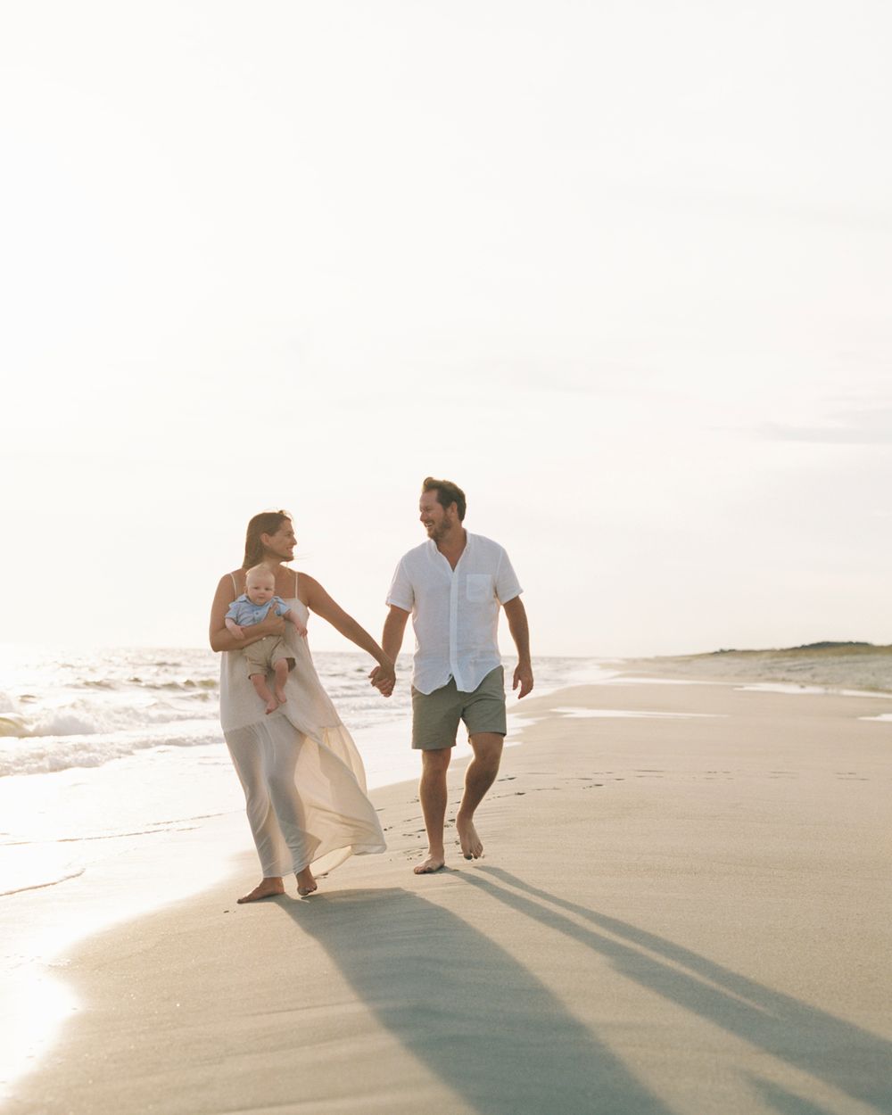 A couple walks together along a sunlit beach shore creating long shadows in the sand during golden hour.