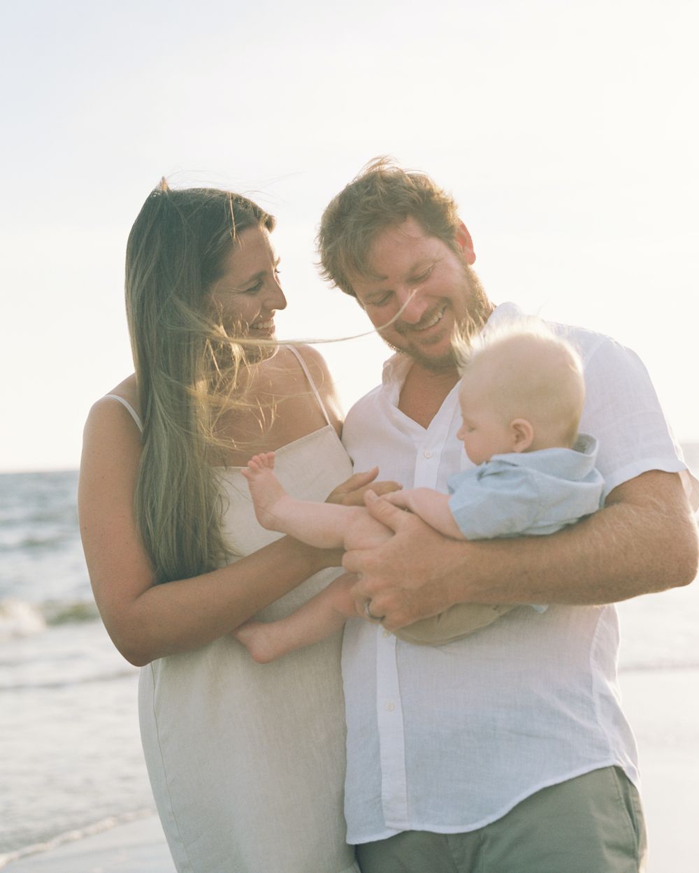 A couple shares tender moments with their baby while standing on a beach at sunset.