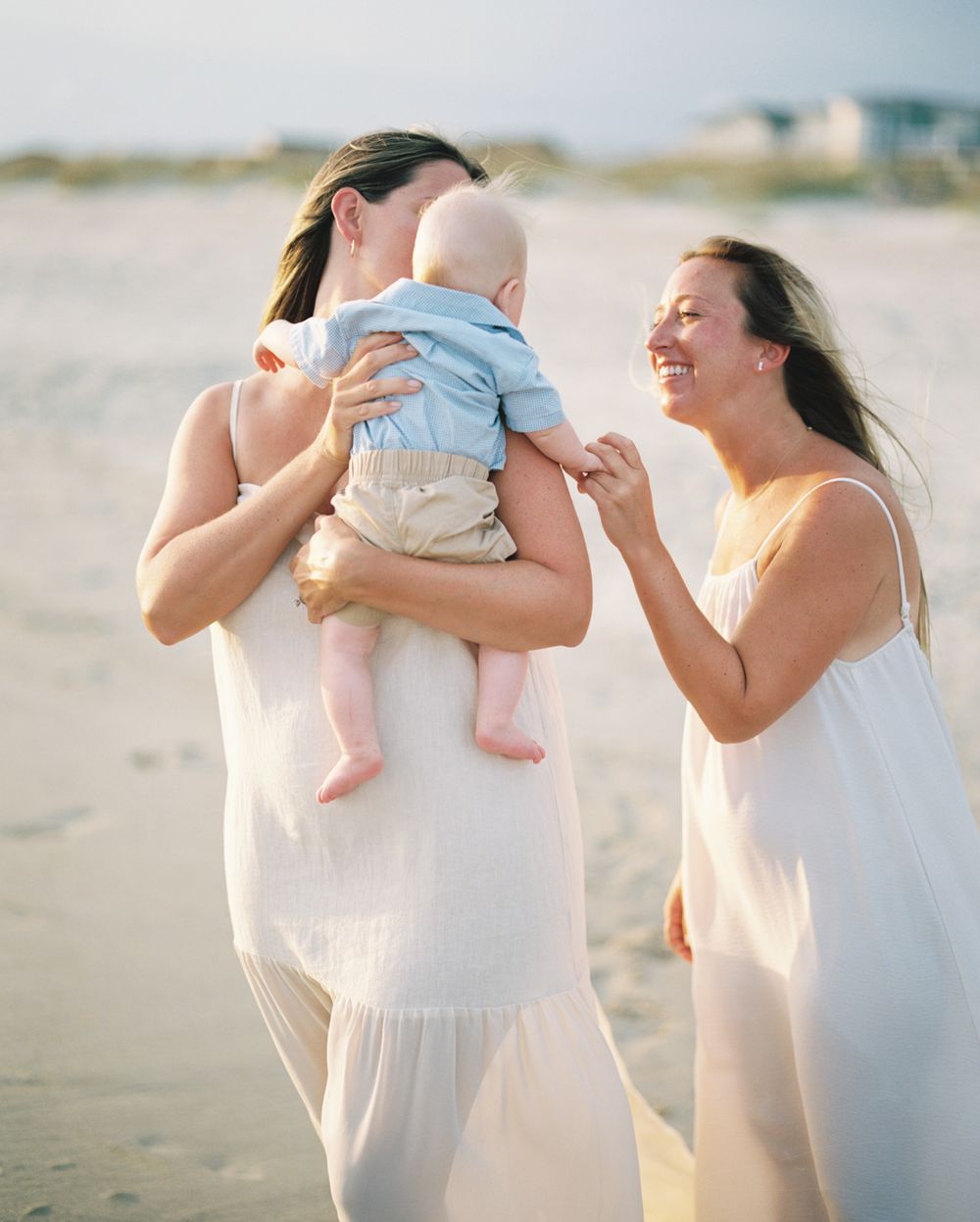 A tender moment on the beach as two people in white dresses share joyful laughter while holding a baby in light blue.