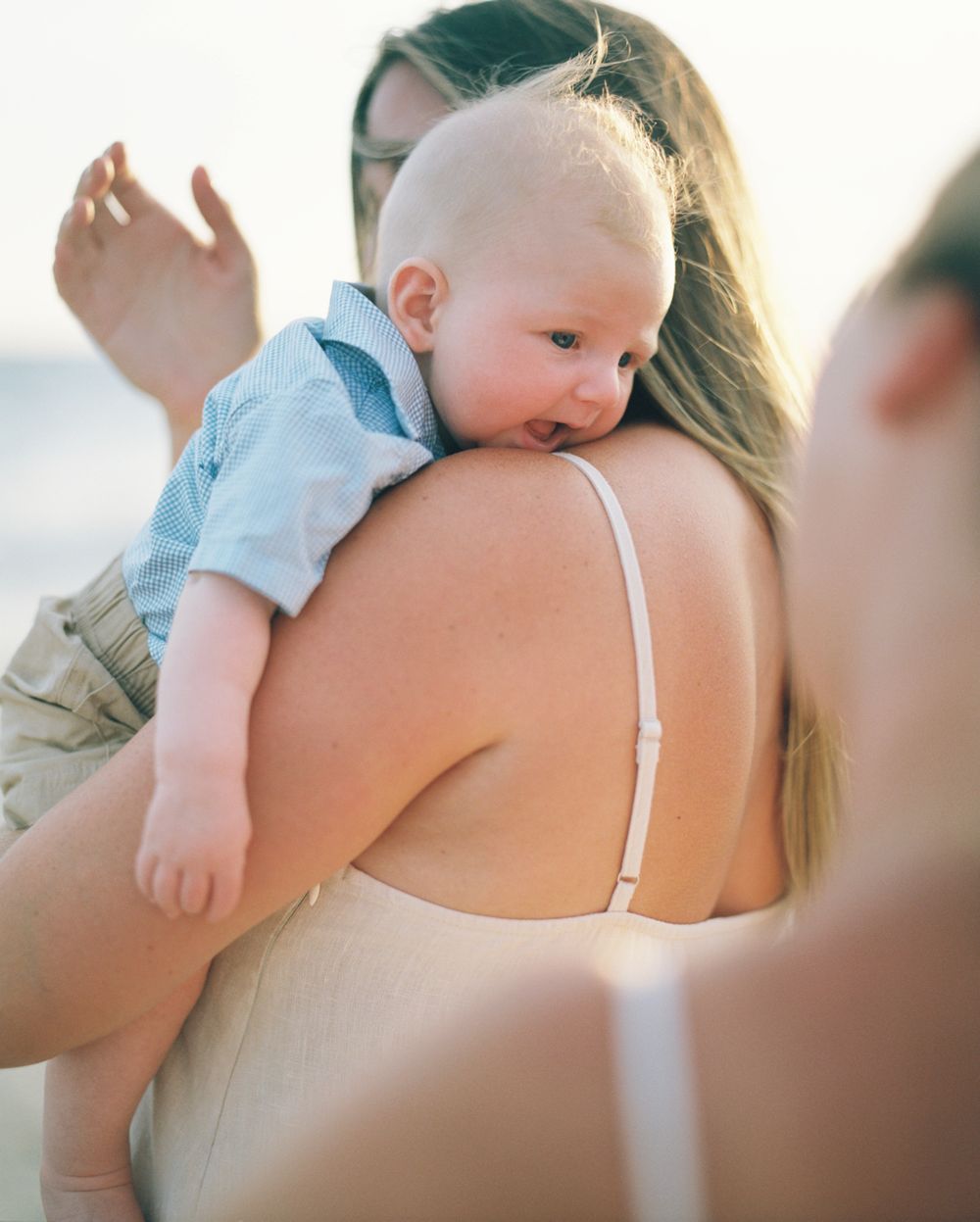 A baby in light blue clothing rests on someone's shoulder against a bright background.