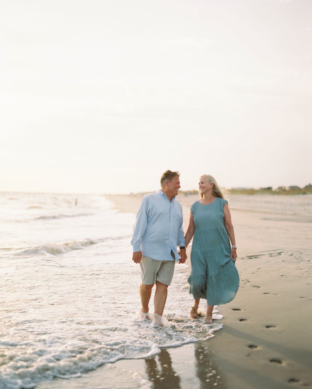 A couple in blue and teal outfits walking together along the beach shoreline at sunset.