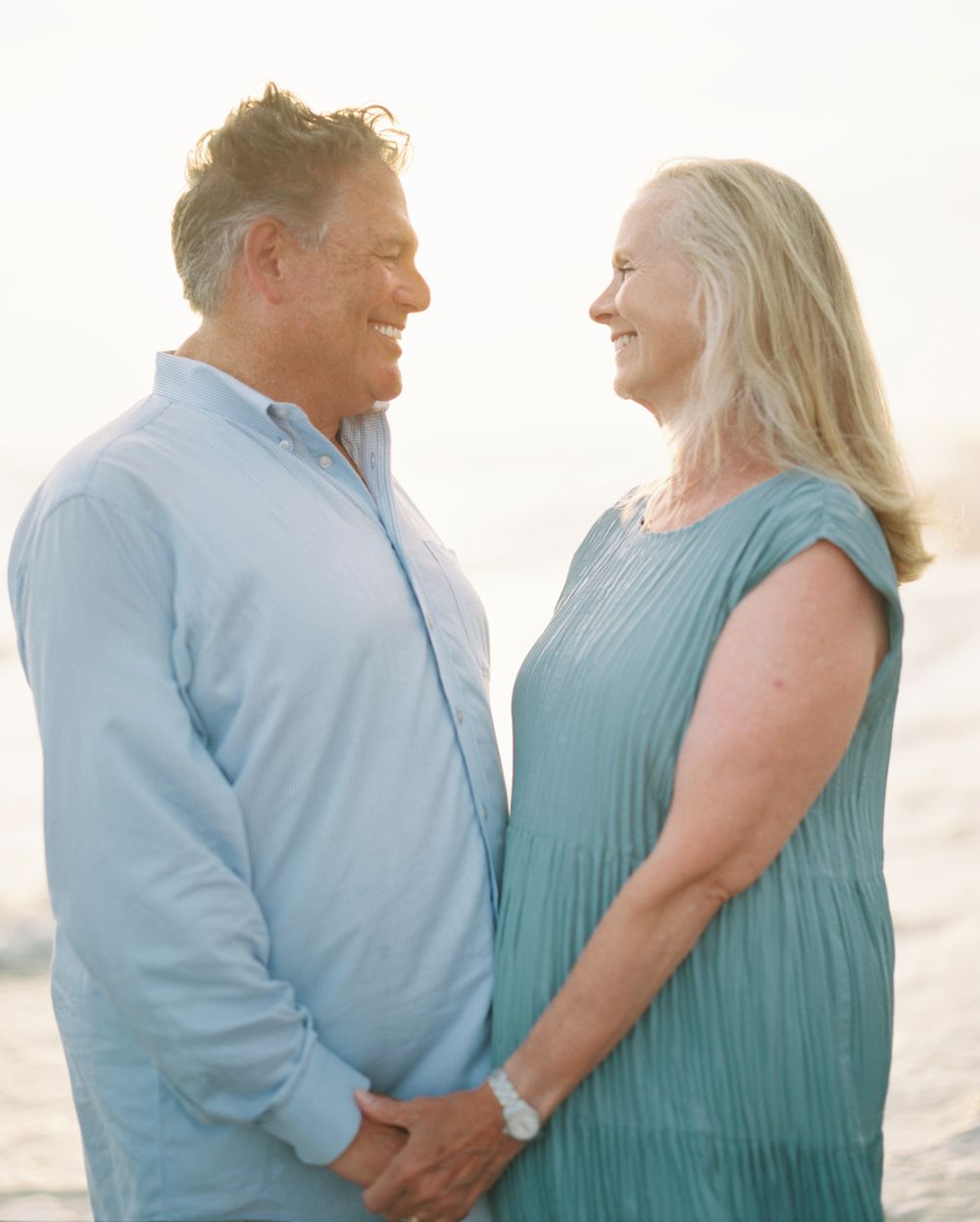 A mature couple wearing light blue and teal clothing share a tender moment together at sunset.