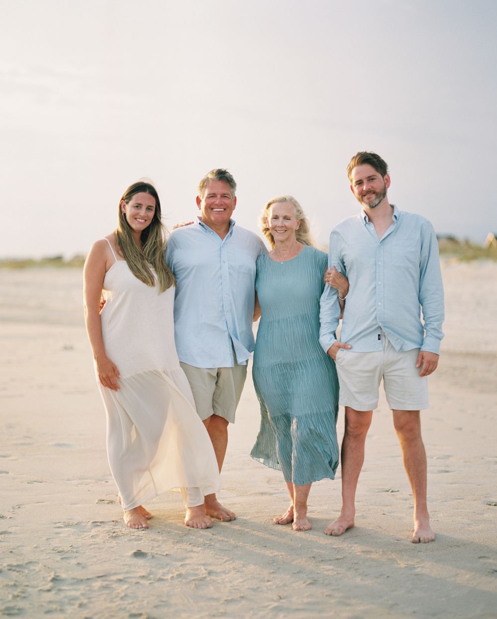 Extended family wearing coordinated blue and white outfits poses casually on a sandy beach.
