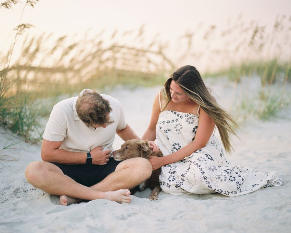 A couple sits on a sandy beach at sunset playing with their small dog while surrounded by sea oats.