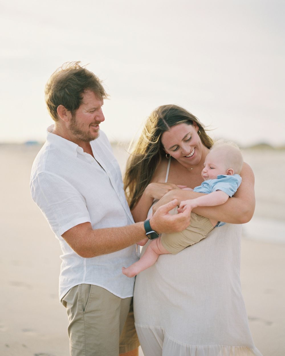 A heartwarming family moment on the beach as parents in light clothing cuddle their infant in the golden light.