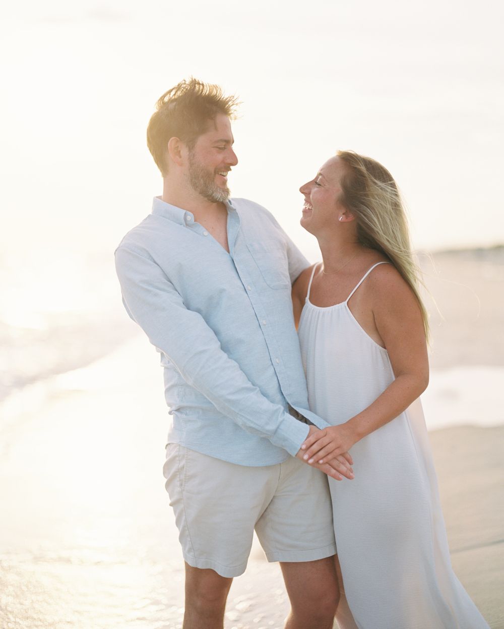A romantic beach photoshoot showing a couple in white and light blue outfits embracing in soft sunlight.