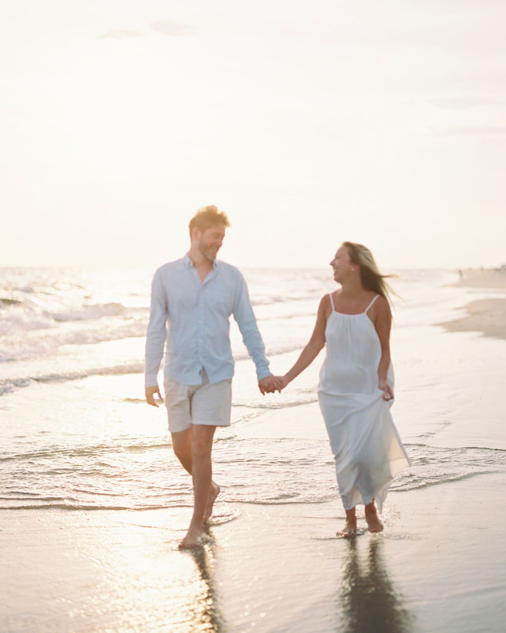 A couple strolls hand in hand along a sunlit beach shoreline in white outfits during golden hour.