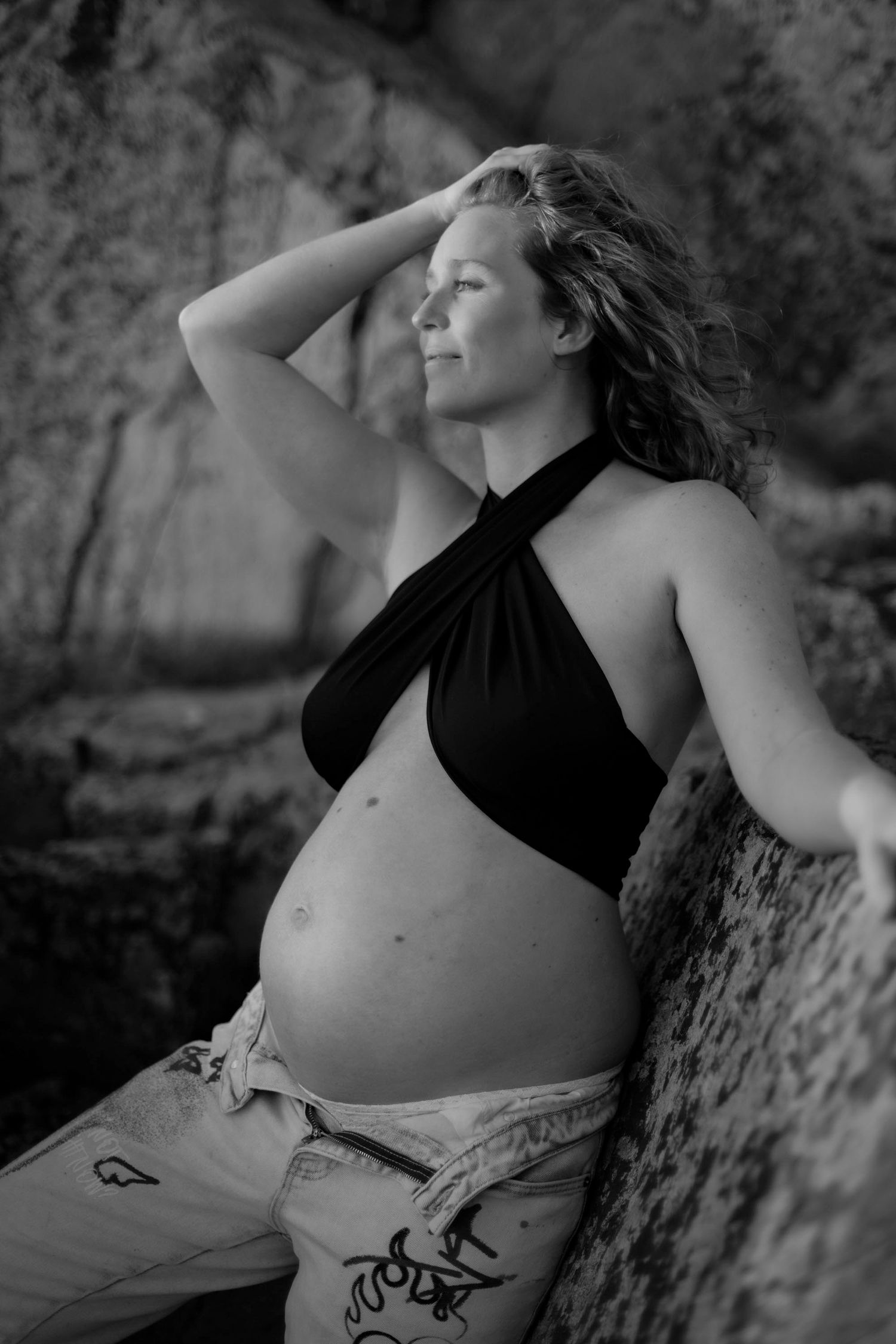 gravid kvinna står lutad mot en klippa. hon har på sig jeans och en svart topp, hon håller handen i håret och bilden är svartvit.
