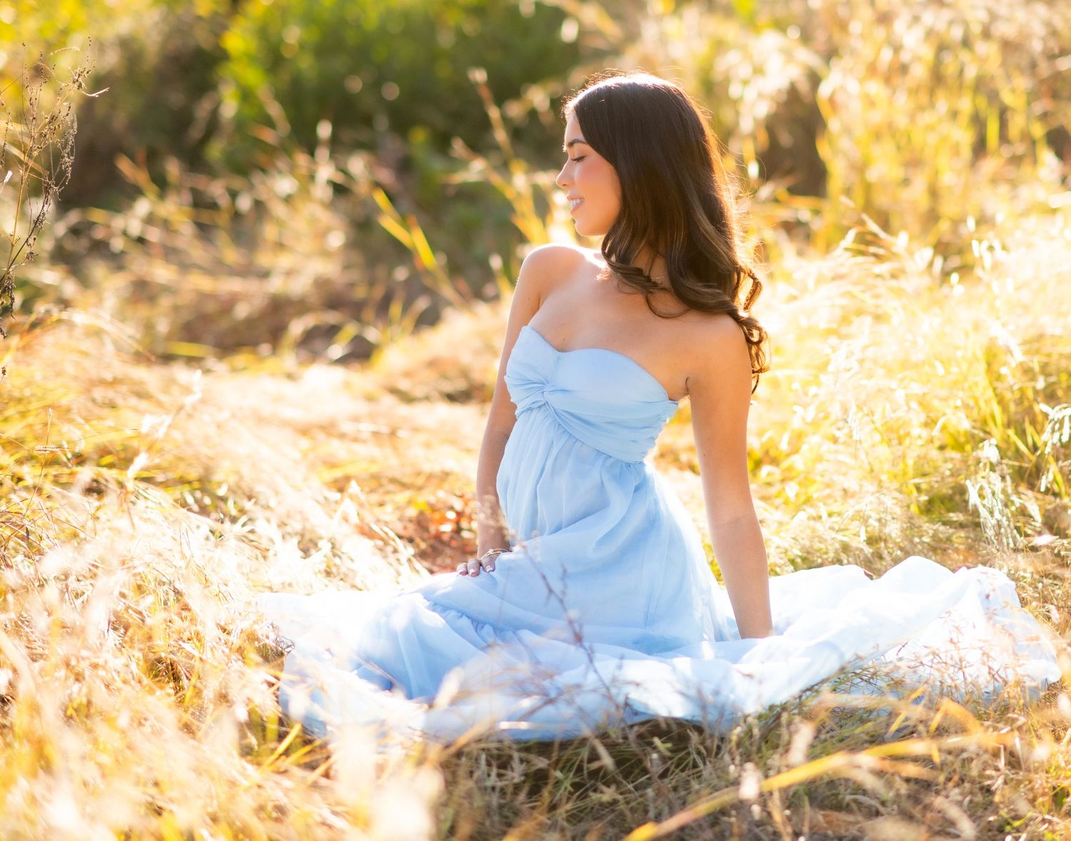 Young woman sitting among tall green grass and wildflowers in golden sunlight, looking serene in a natural outdoor setting.