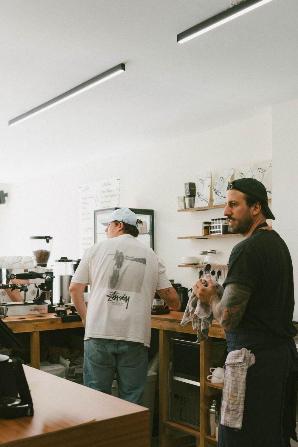 Deux baristas travaillant dans un café minimaliste moderne, un homme tatoué en noir tenant une tasse.