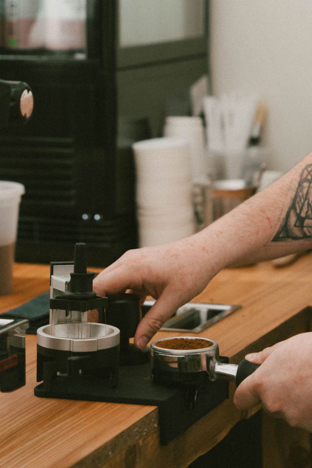 Barista tatoué tamponnant du marc d'espresso frais dans un porte-filtre sur un comptoir de bar à café en bois.