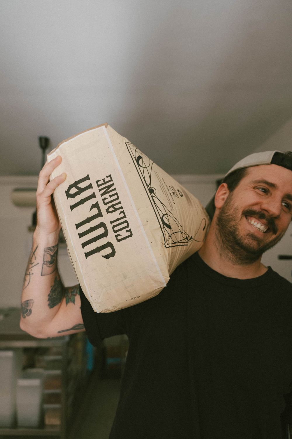 Un homme tatoué souriant tenant un sac de farine sur son épaule dans un cadre intérieur décontracté. Photographe var café restaurants