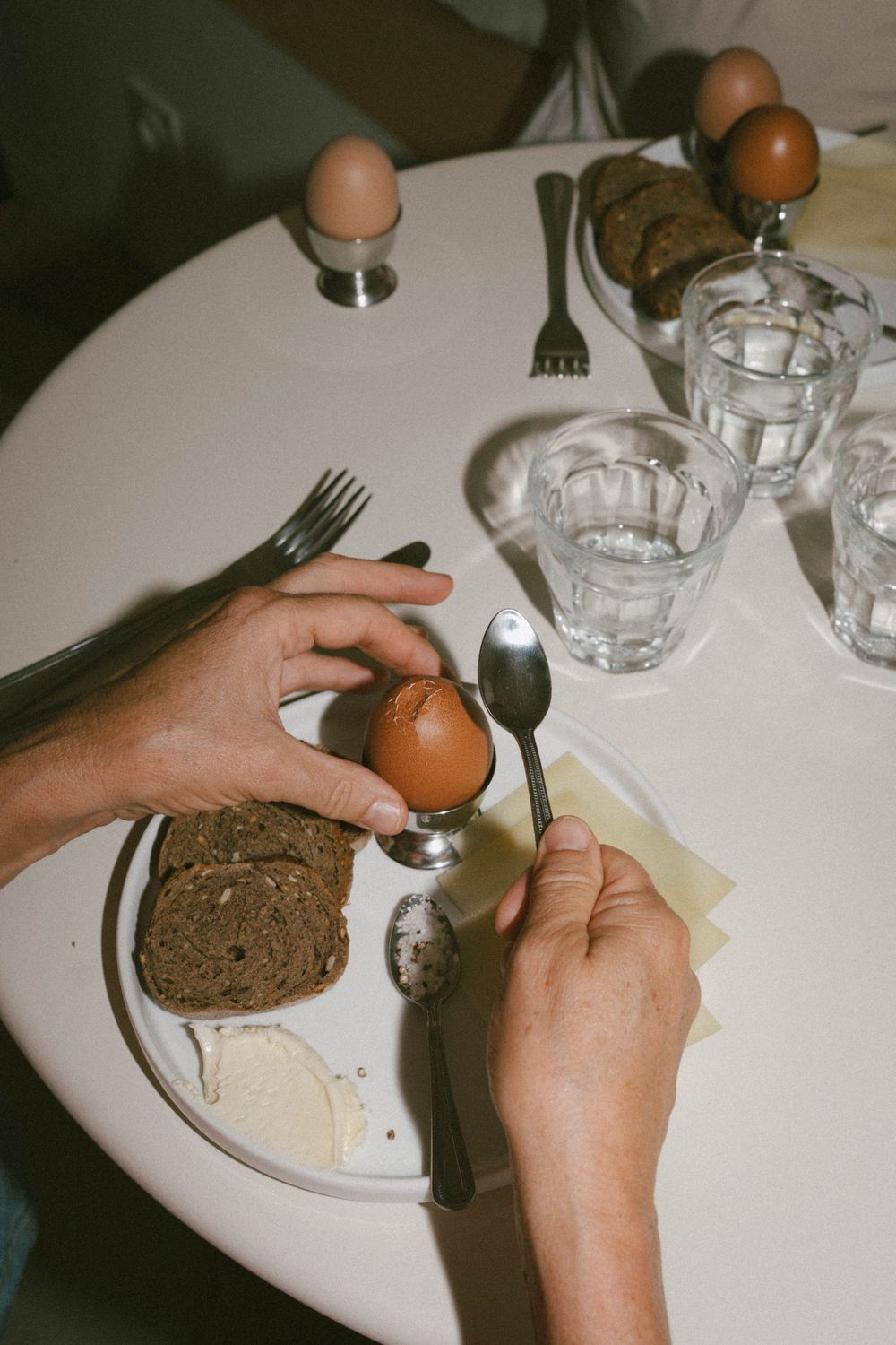Les mains cassent un œuf à la coque dans une tasse en argent, avec du pain de seigle, du beurre, des verres d'eau sur une table ronde