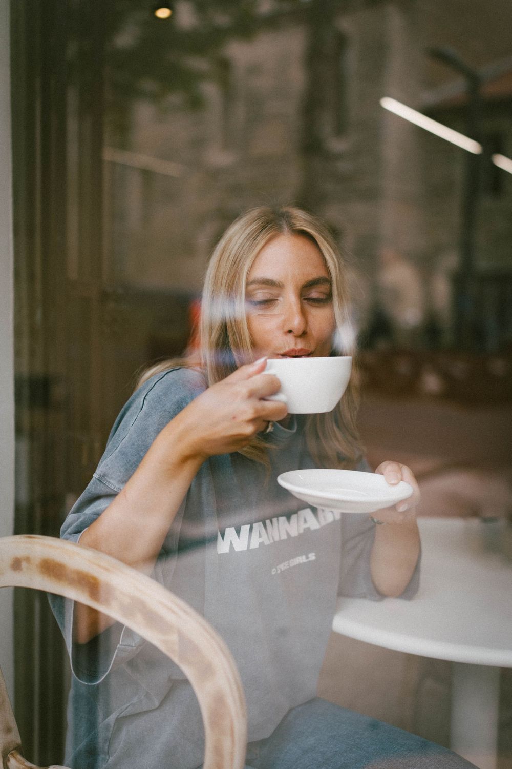 Woman sipping coffee from a white cup at a cafe table, photographed through a glass window.