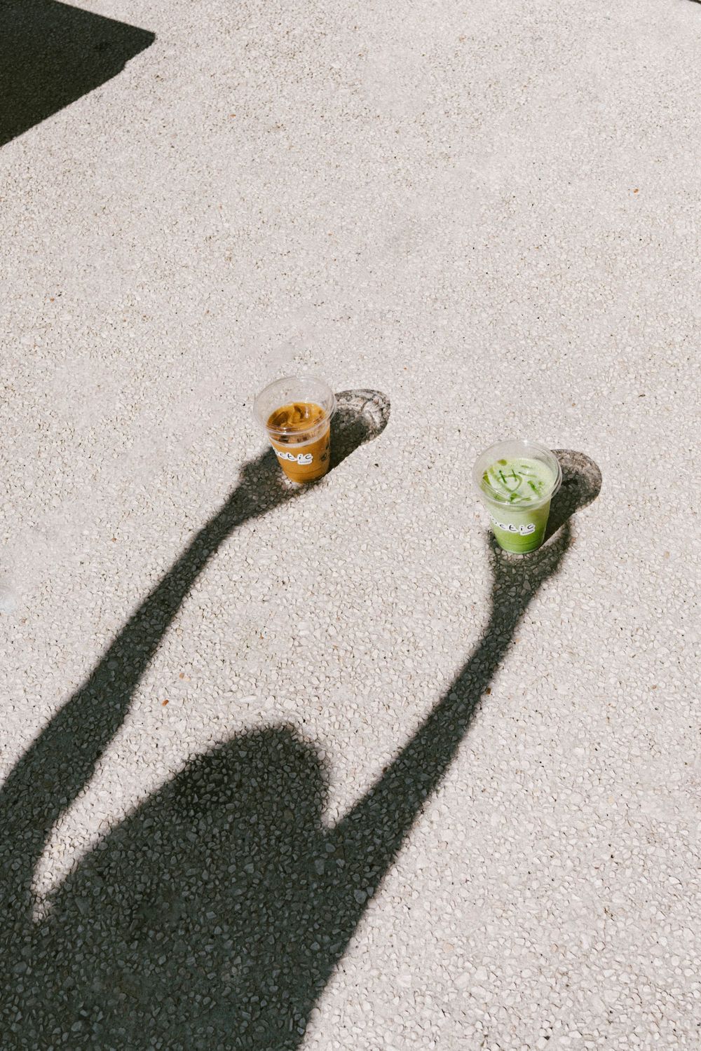 Deux boissons glacées sur un trottoir ensoleillé projetant de longues ombres, un café ambré et une boisson matcha verte. Photographe var