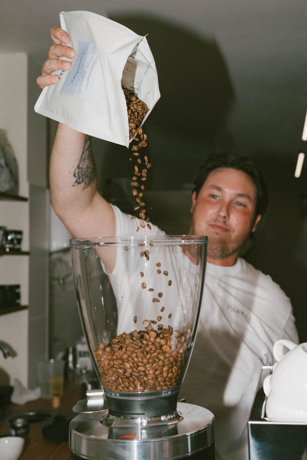 Barista pouring fresh coffee beans from a white bag into a commercial coffee grinder in a cafe.