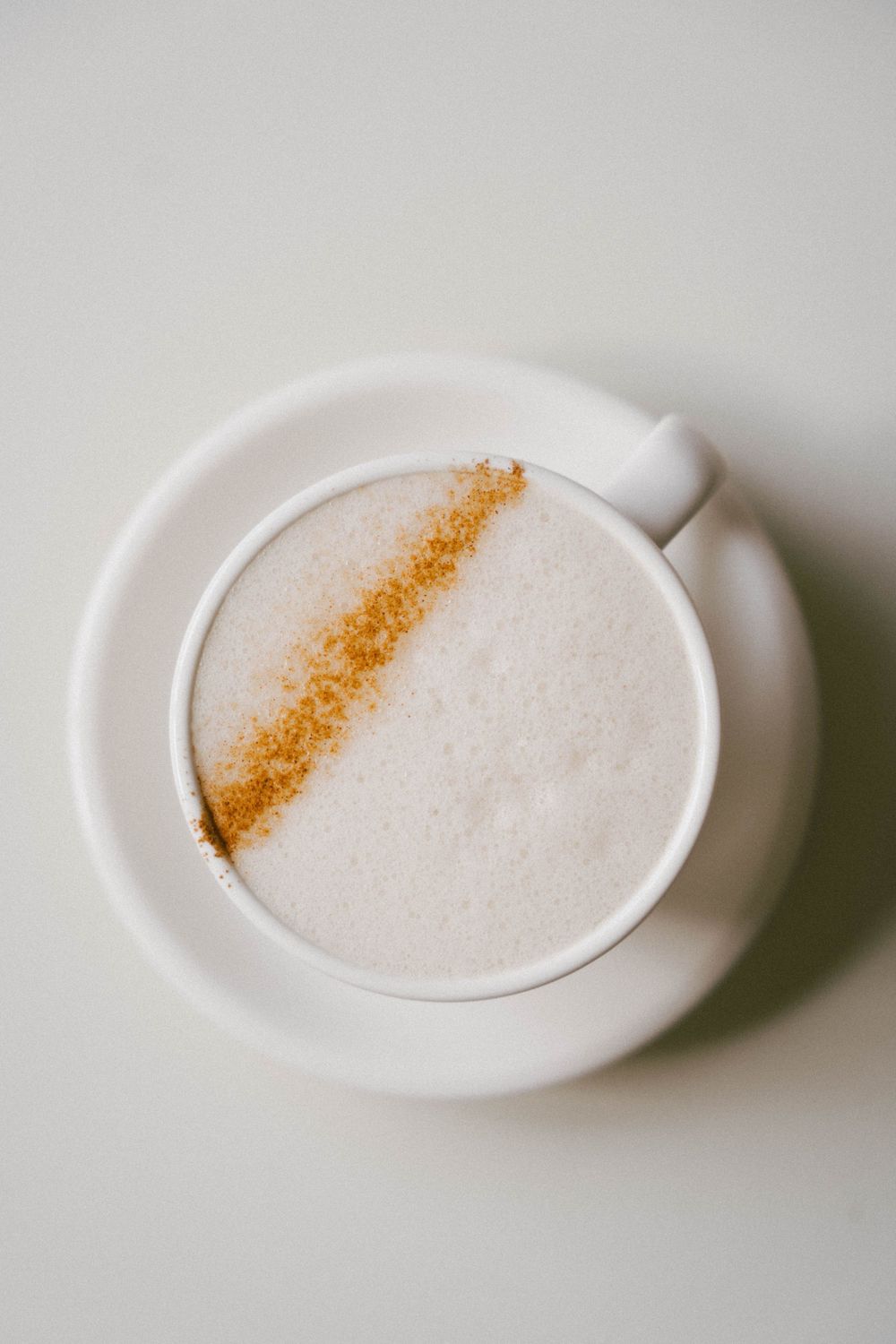 Vue aérienne d'une tasse en céramique blanche de café au lait mousseux avec un saupoudrage diagonal de cannelle sur une soucoupe blanche.