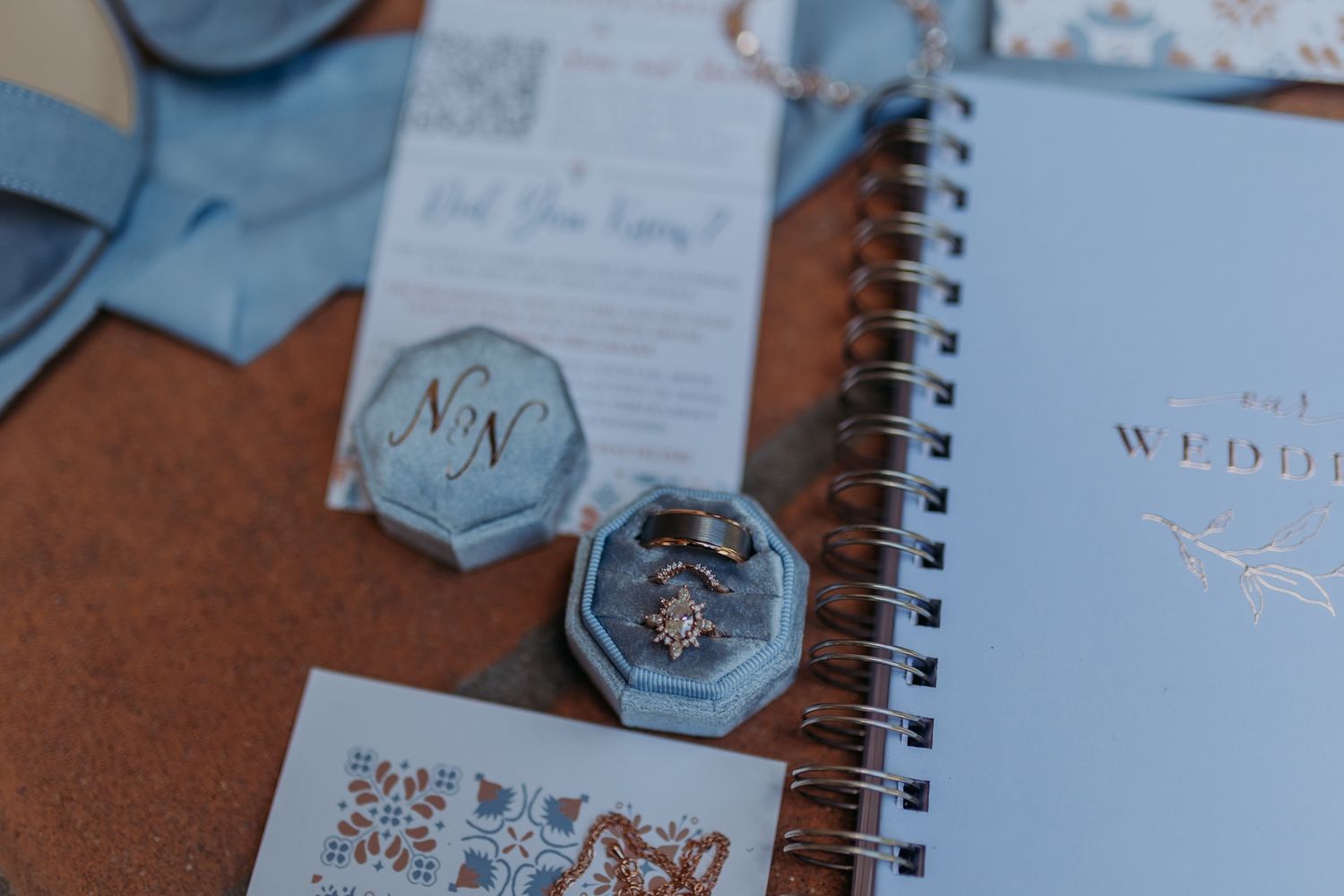 Blue velvet ring box with diamond rings beside a wedding planner notebook and elegant blue stationery suite.