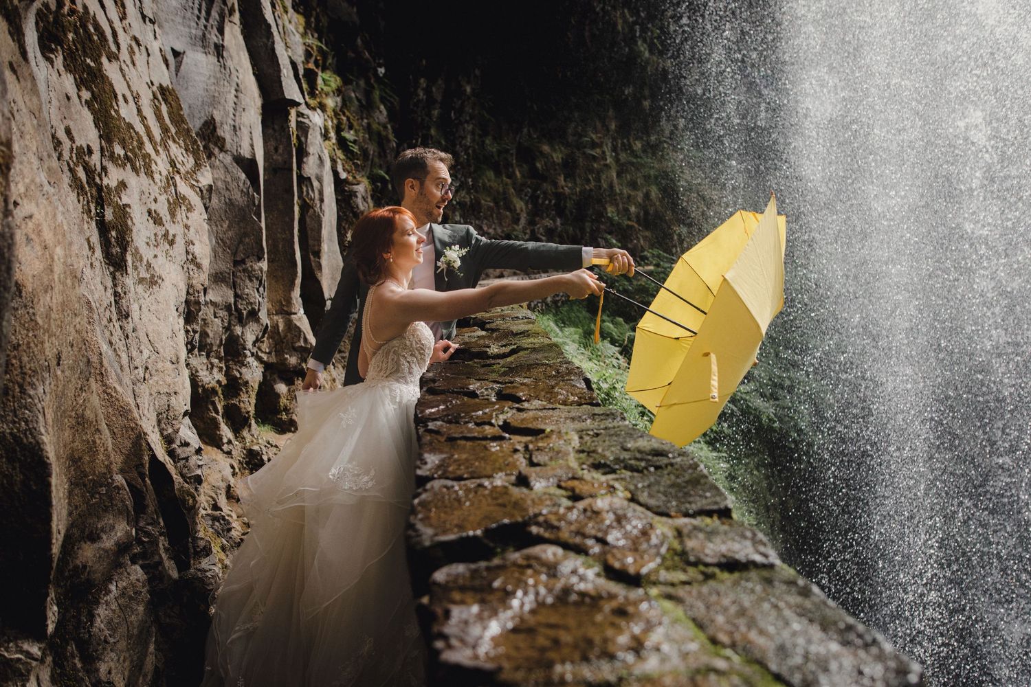 Wedding party of six smiling in front of a waterfall, bridesmaids in teal dresses, groomsmen in tan suits.