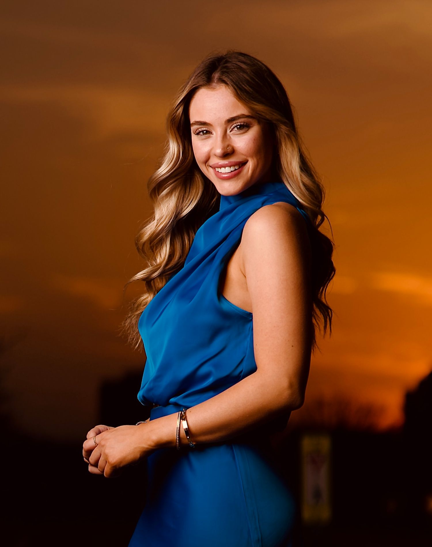 Woman in blue sleeveless dress poses against warm sunset sky in artistic portrait series.