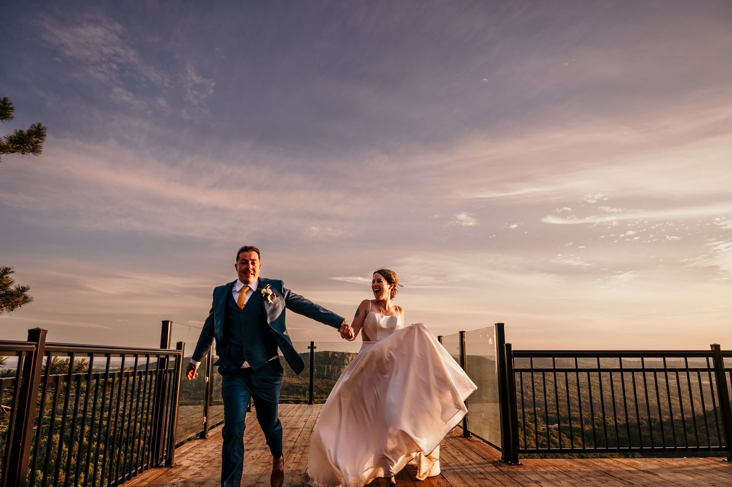 A couple in wedding attire embraces at sunset on a balcony overlooking a scenic mountain landscape.