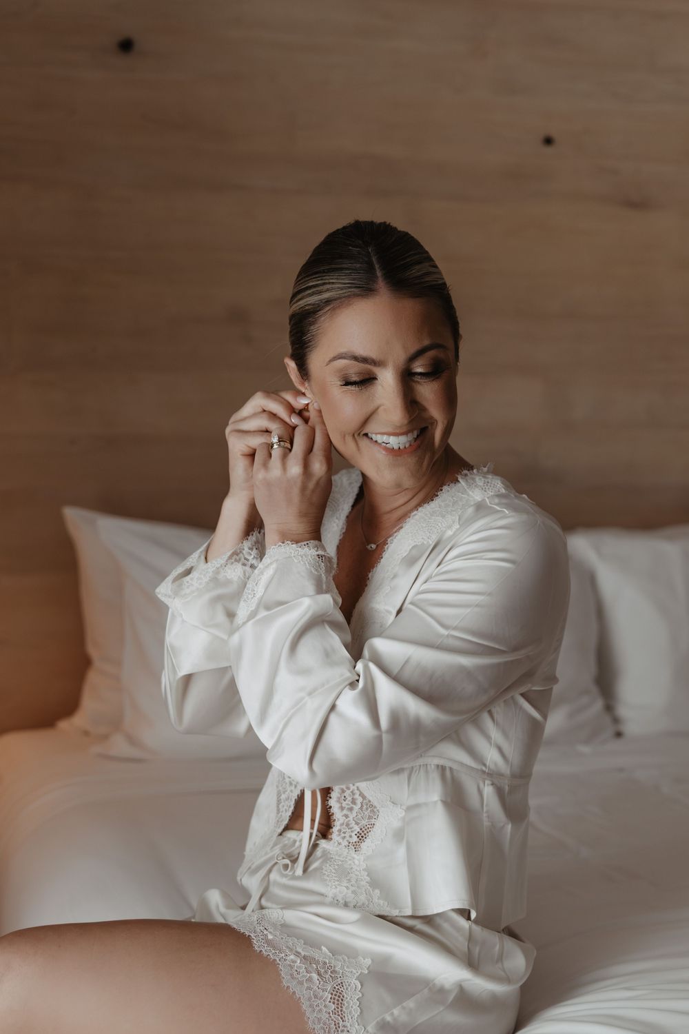 A person wearing a white silk robe sits on a bed smiling while putting on earrings in warm lighting.