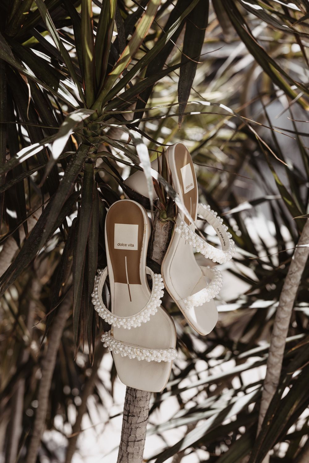 White decorative wedding shoes hanging on tree branches with lace details against a natural outdoor background.