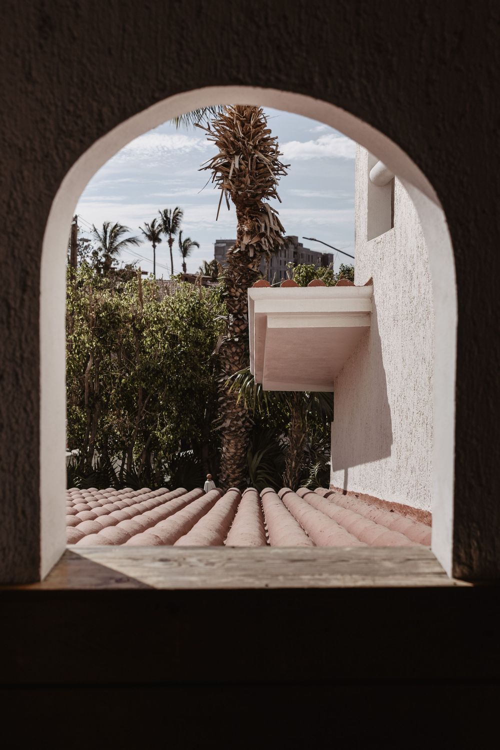 A series of arched windows frame a Mediterranean garden with palm trees and white stucco walls against a cloudy sky.