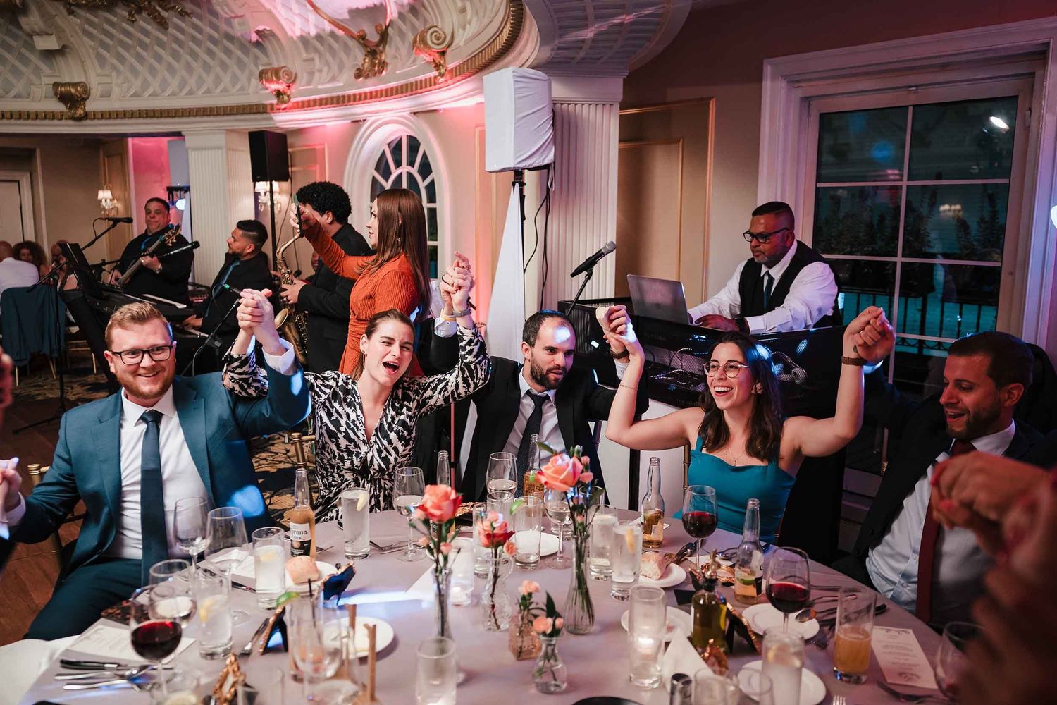 Wedding guests raising their hands and cheering at a reception table while the live band performs at the Lenox Hotel Boston.