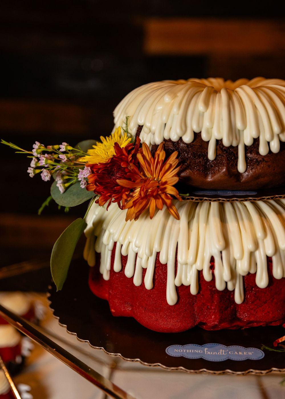 Two-tier red velvet bundt cake with cream cheese frosting drizzle, topped with fall flowers including sunflowers and mums.