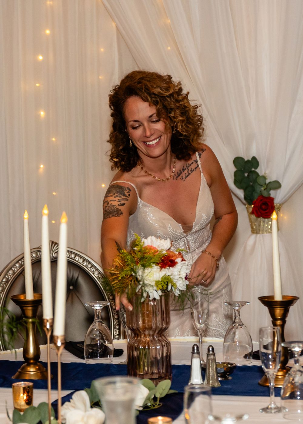 Smiling bride with tattoos arranging colorful floral centerpiece at elegant candlelit wedding reception table at a Venue on the Ridge