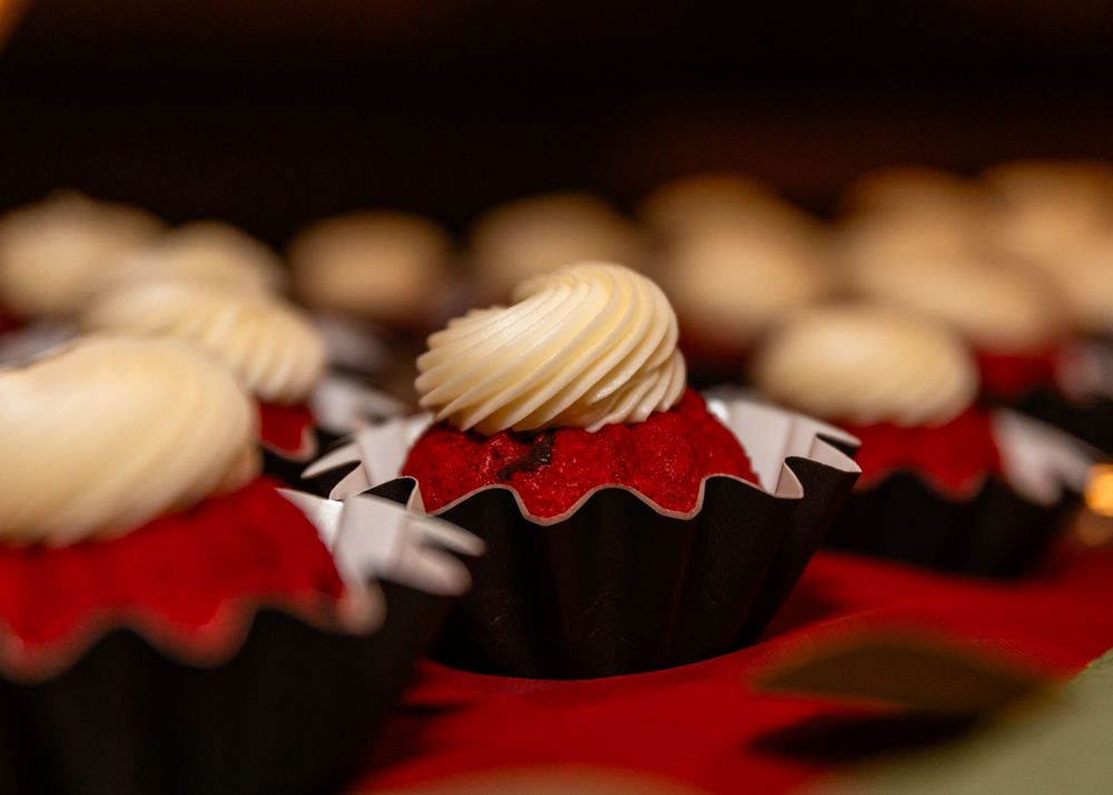 Red velvet cupcakes with creamy white frosting swirls in dark chocolate wrappers displayed on a red surface.