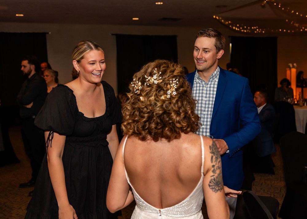 Bride in backless wedding dress chats with guests in black dress and blue suit at elegant wedding reception at a Venue on the Ridge