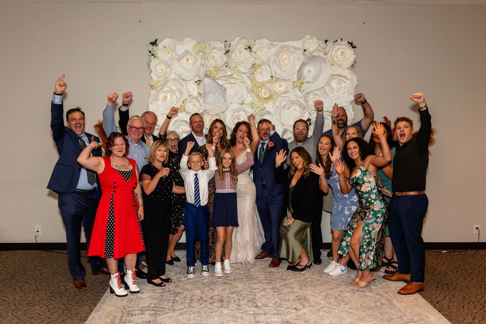 Large wedding group celebrating in front of a white floral wall backdrop, raising hands in joy at a Venue on the Ridge in Elkhorn NE