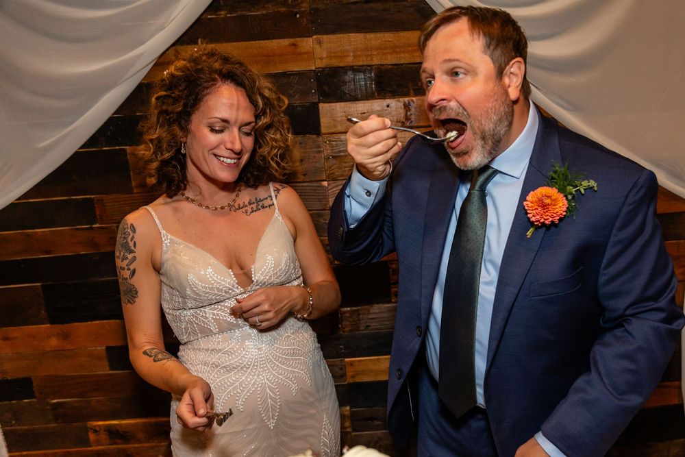 Bride & groom laughing while cutting wedding cake, bride in lace dress with tattoos, groom in blue suit at a Venue on the Ridge in Elkhorn