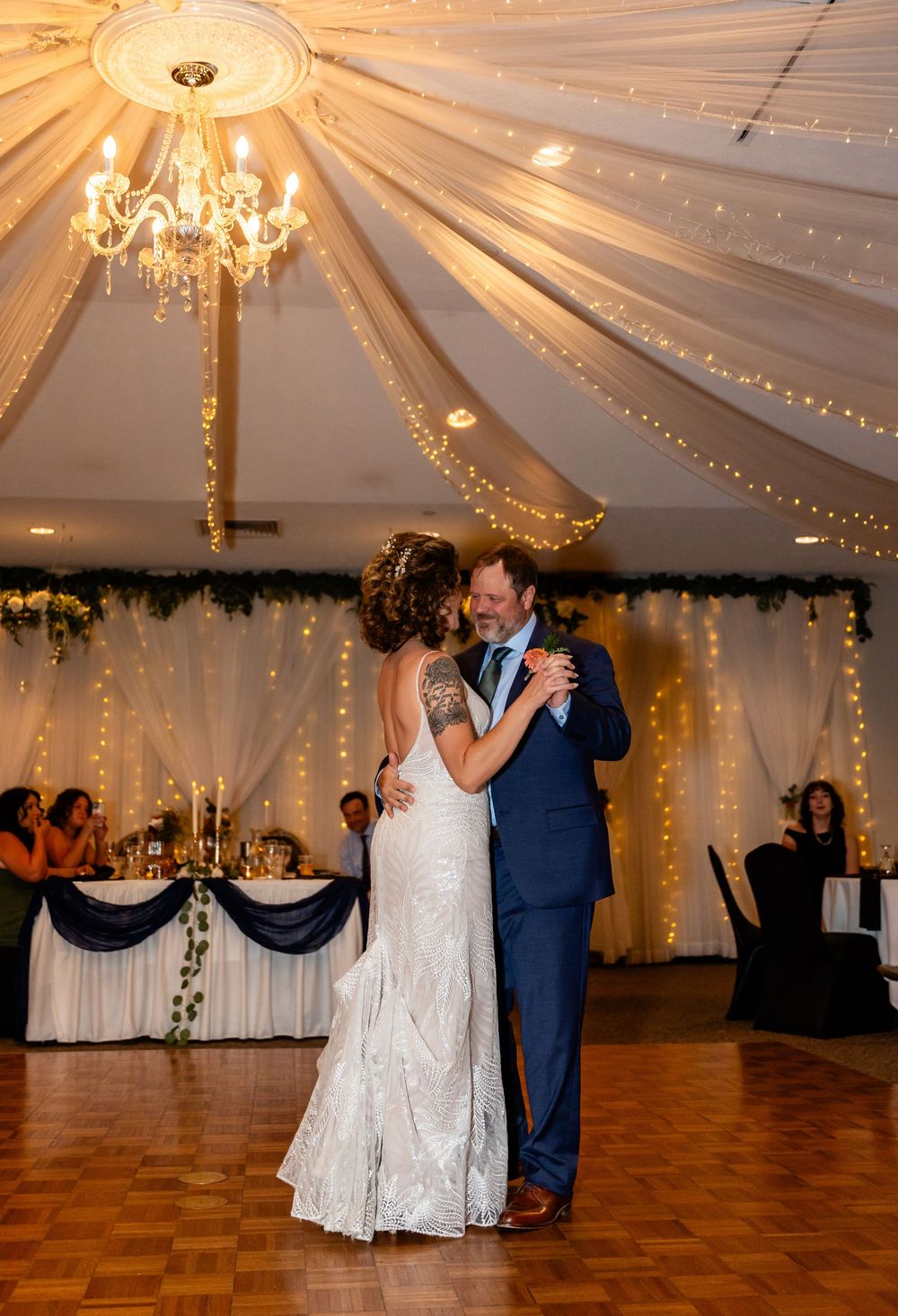 Bride and groom share their first dance under a chandelier with white draping and fairy lights at elegant wedding reception.
