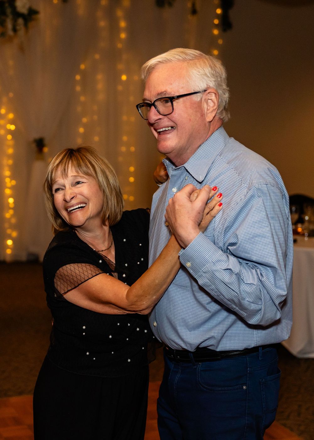 Elderly couple dancing and smiling at a formal event with warm fairy lights in the background.